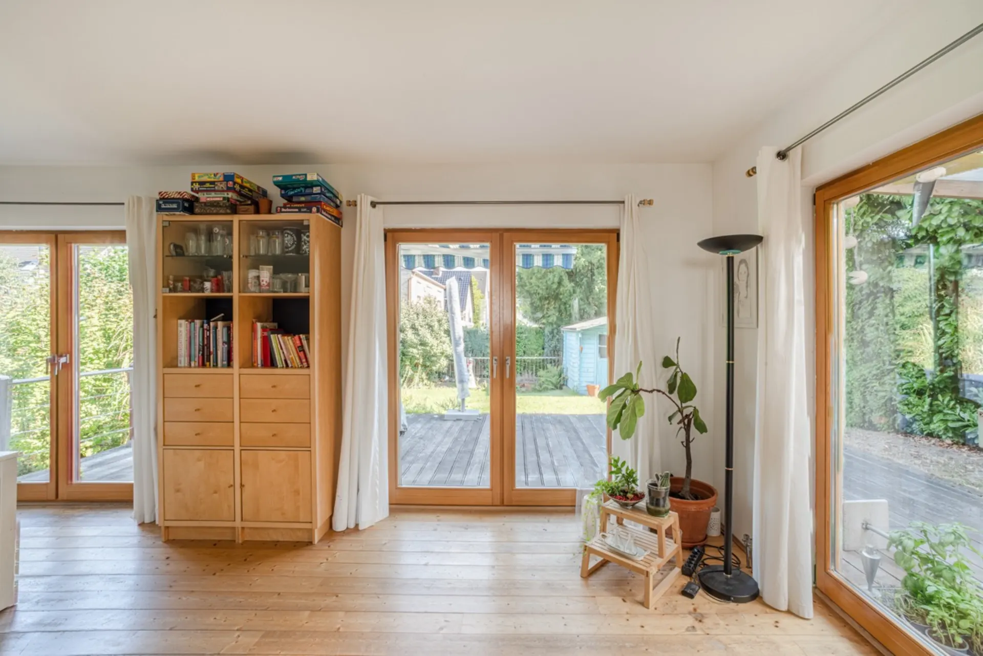 Wohnzimmer mit Holzboden, großen Fenstern, Regal, Zimmerpflanzen und Blick auf Terrasse und Garten.