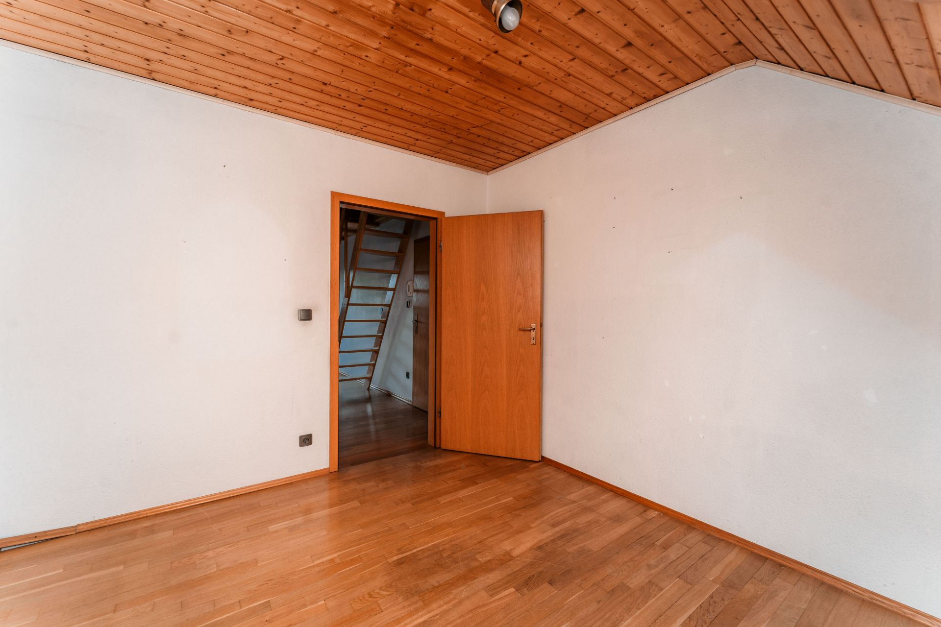 Leeres Zimmer mit Holzdecke, Holzboden und geöffneter Tür, dahinter eine Treppe sichtbar.