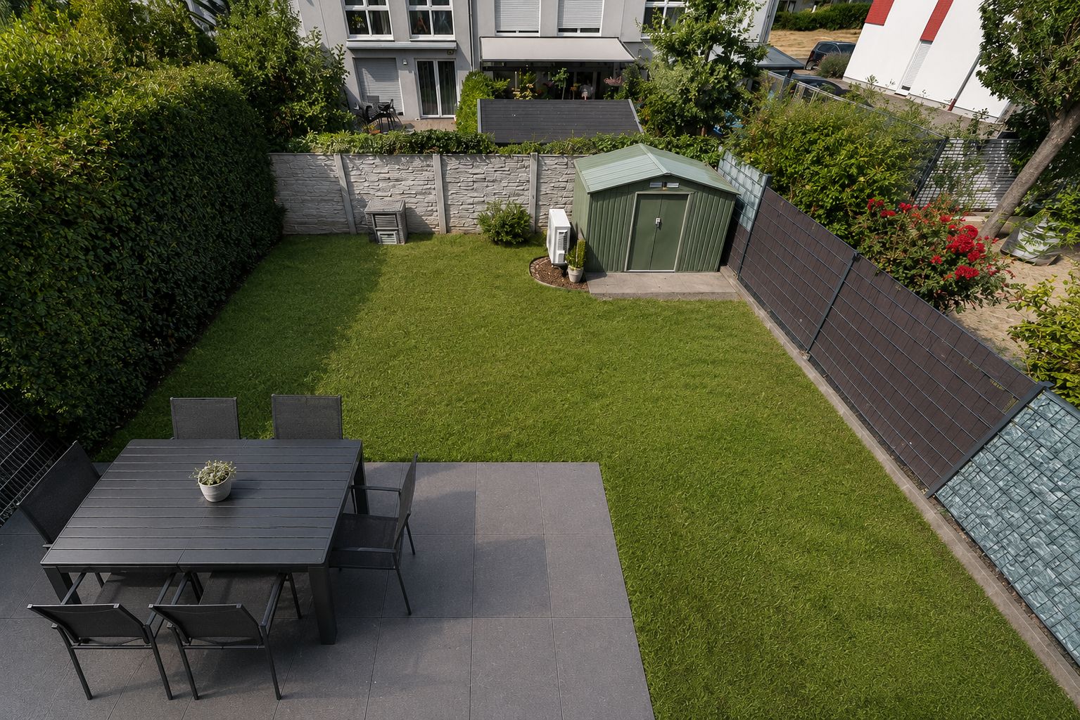 Gepflegter Garten mit Terrasse, Esstisch, Stühlen und grünem Geräteschuppen.