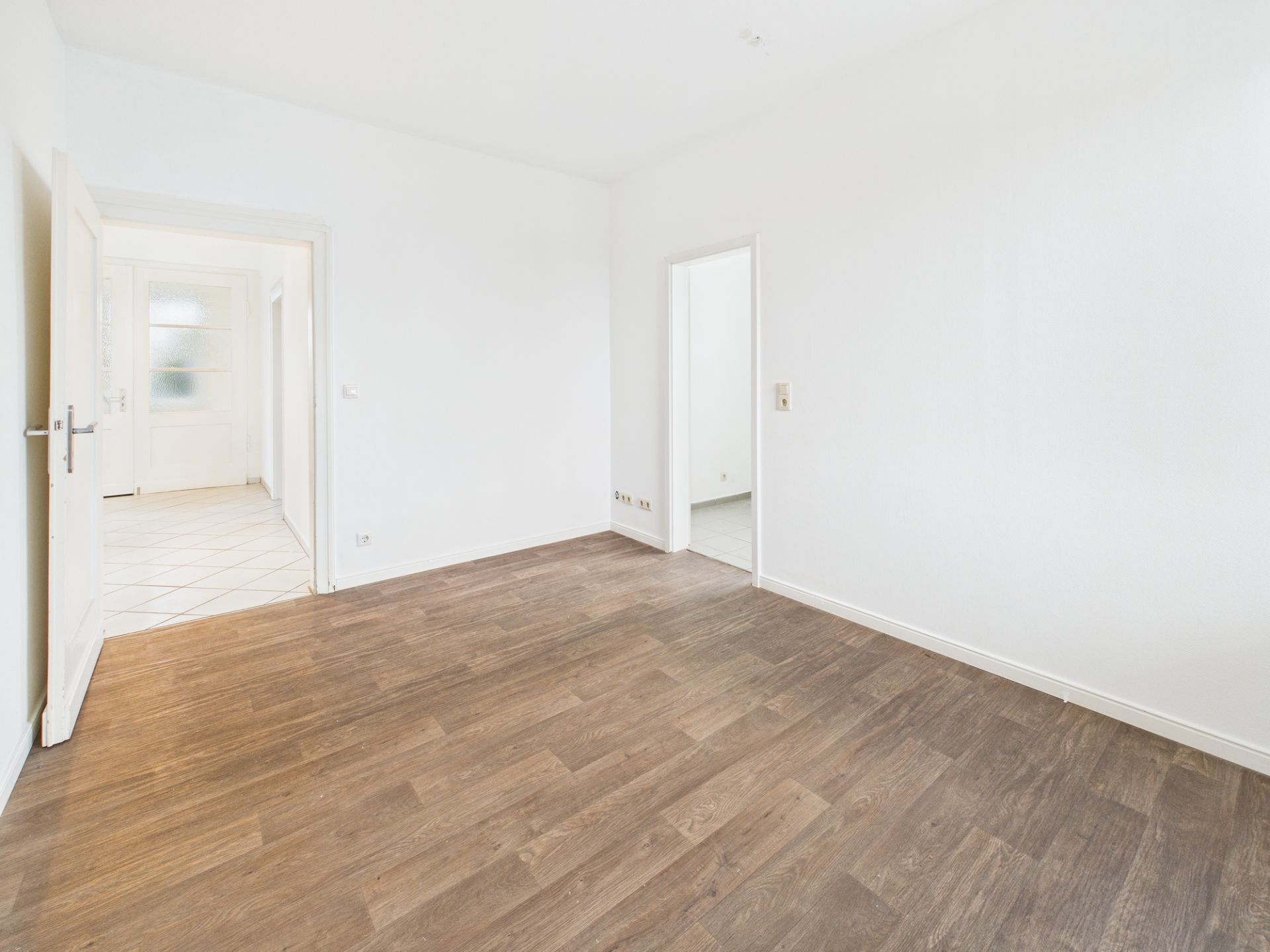 Leerer Raum mit dunklem Holzboden und weißen Wänden, zwei Türen führen in andere Zimmer.