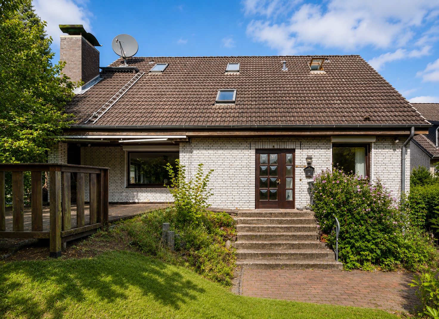 Haus mit braunem Satteldach, weißen Klinkersteinen und Treppe zum Eingang, umgeben von Garten.