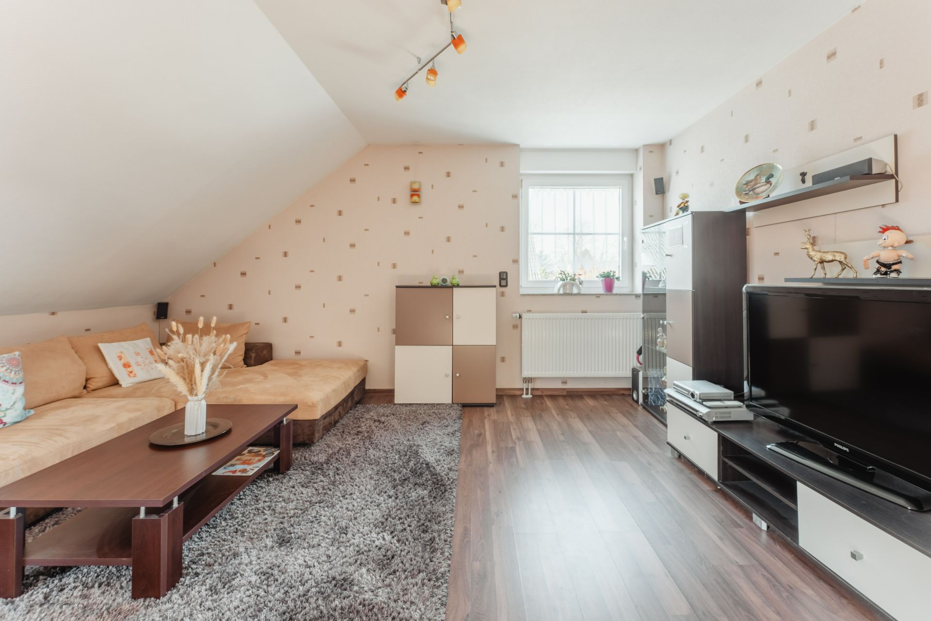 Wohnzimmer mit Ecksofa, Teppich, TV-Schrank, Regal und Fenster mit Tageslicht.