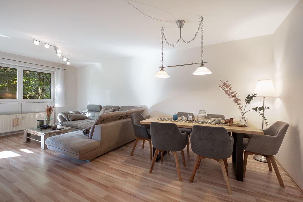 Helles Wohnzimmer mit grauem Sofa, Holztisch und sechs Stühlen, Lampe über dem Esstisch.