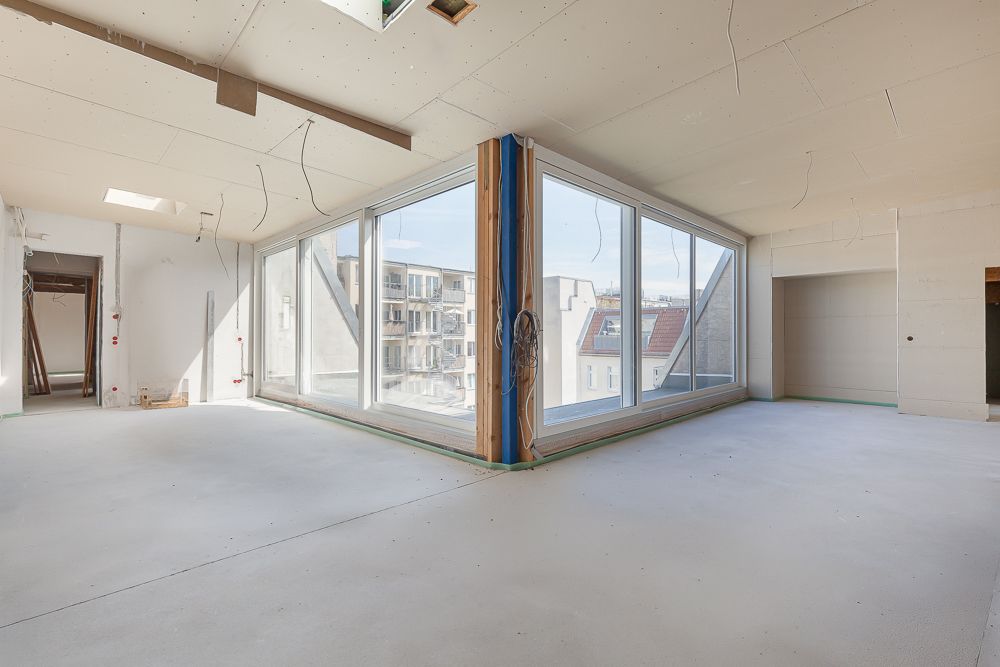 Heller, unfertiger Innenraum mit großen Fenstern und Blick auf benachbarte Gebäude.