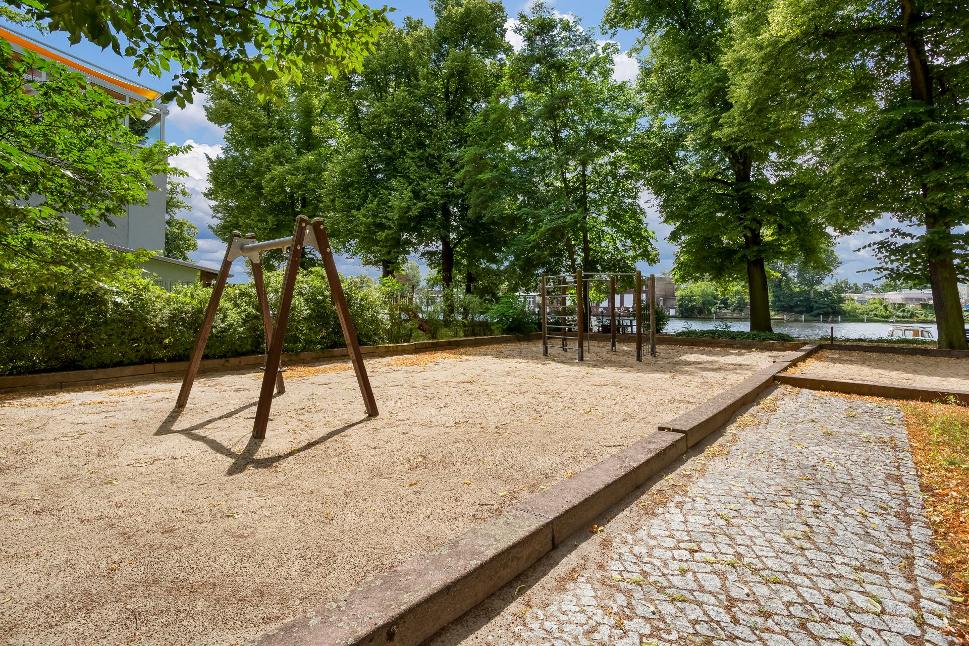 Ein Spielplatz mit Schaukel und Klettergerüst auf Sand, umgeben von Bäumen und Gehweg.