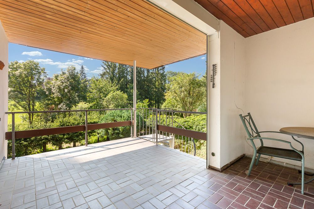 Überdachter Balkon mit Fliesenboden, Tisch und Stuhl, Blick auf grüne Bäume.