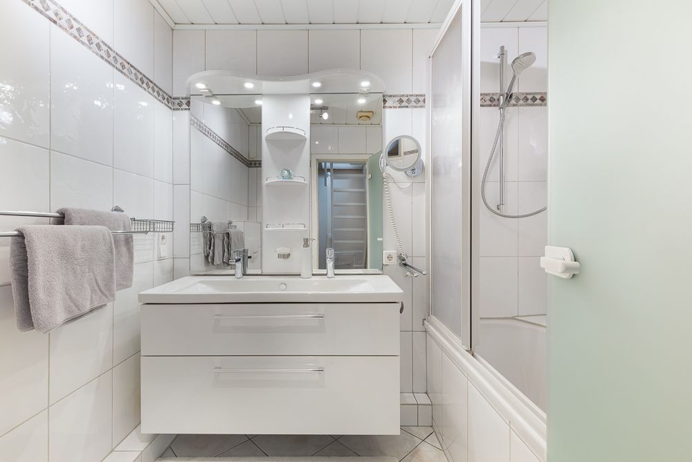 Helles Badezimmer mit Waschbecken, Spiegel, Handtüchern und Duschwanne mit Glasabtrennung.