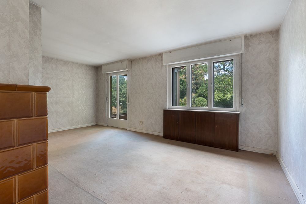 Leeres Wohnzimmer mit großem Fenster, Tür zum Garten und braunem Kamin links.
