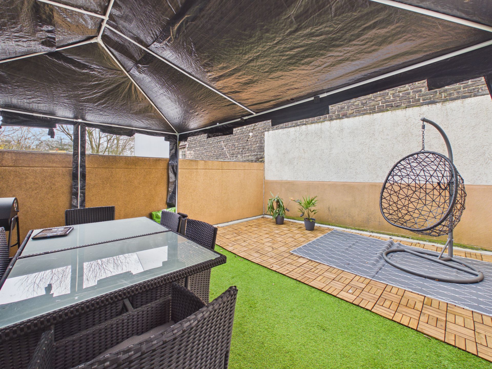Überdachte Terrasse mit Esstisch, Stühlen, Hängesessel und zwei Pflanzen auf Kunstrasen.