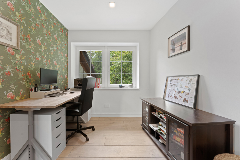 Helles Arbeitszimmer mit Schreibtisch, Stuhl, Fenster und Blumen-Tapete an einer Wand.