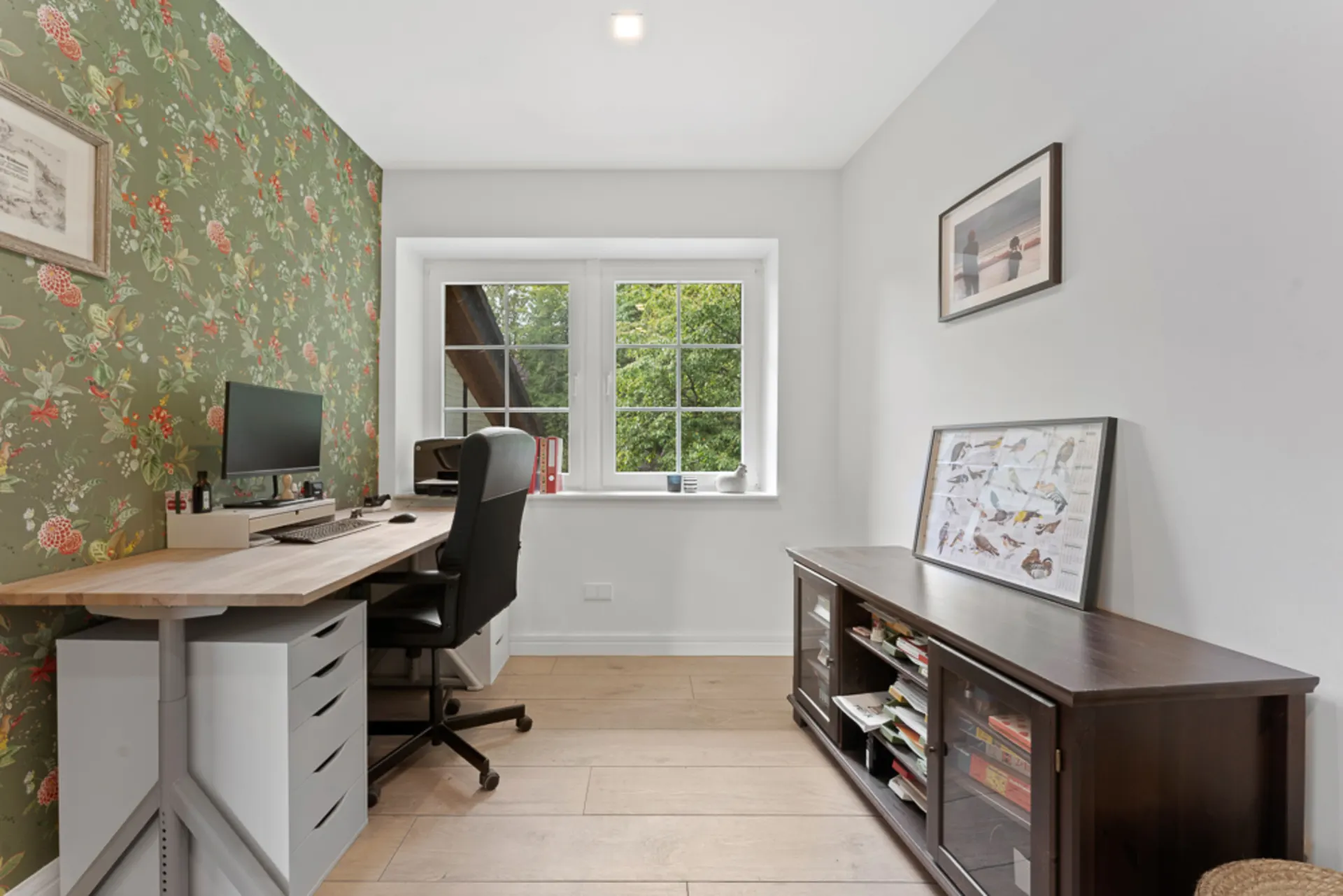 Helles Arbeitszimmer mit Schreibtisch, Stuhl, Fenster und Blumen-Tapete an einer Wand.
