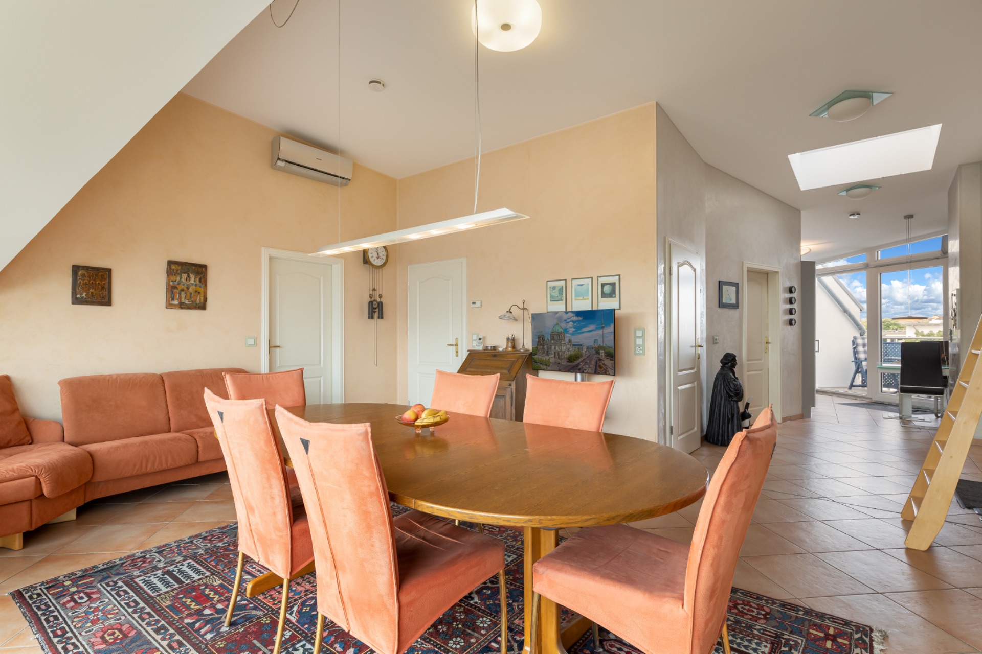 Helles Esszimmer mit rundem Holztisch, sechs Stühlen, Sofa und Teppich auf Fliesenboden.