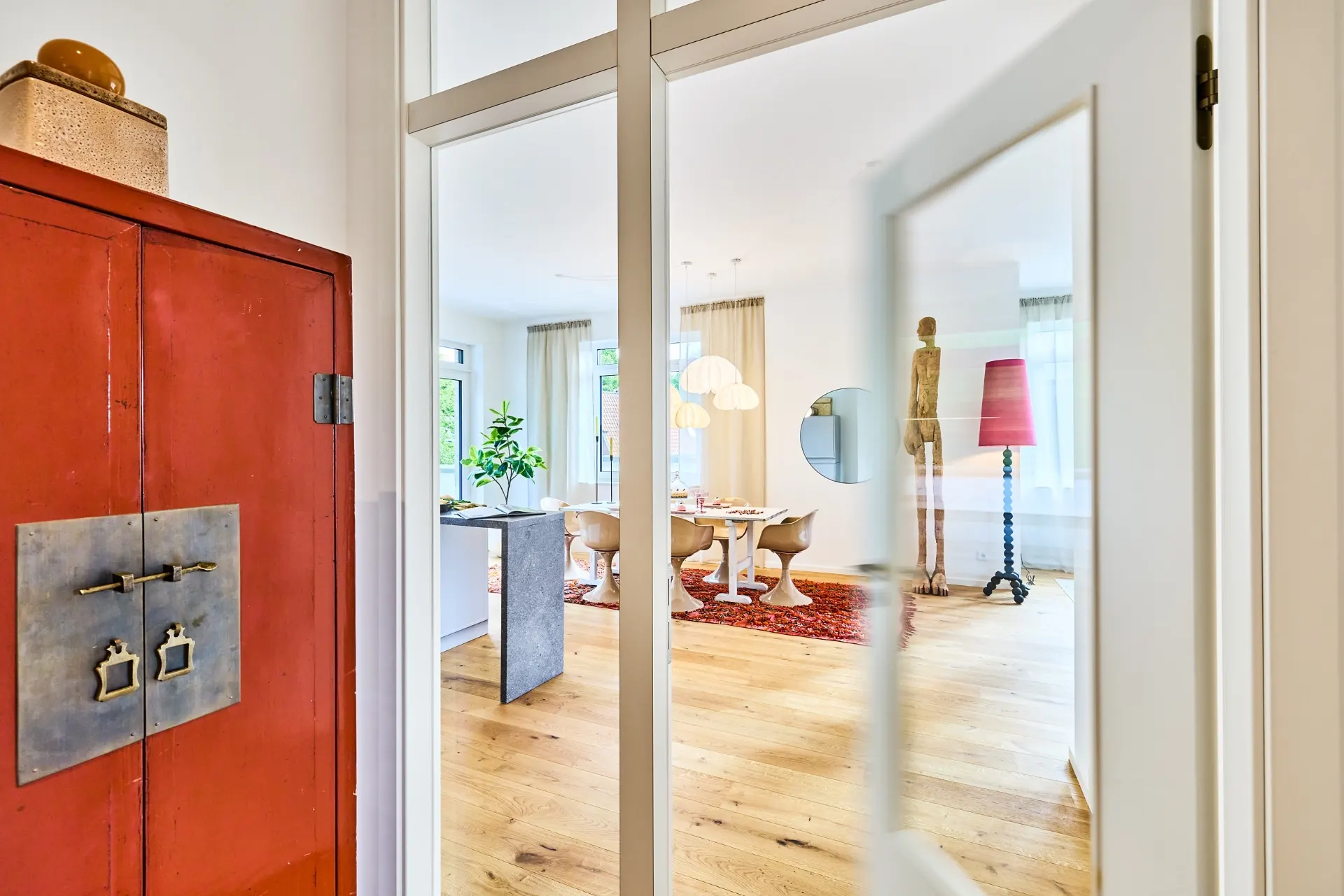 Blick durch Glastür auf helles Wohnzimmer mit Holzboden, Esstisch und rotem Schrank im Vordergrund.