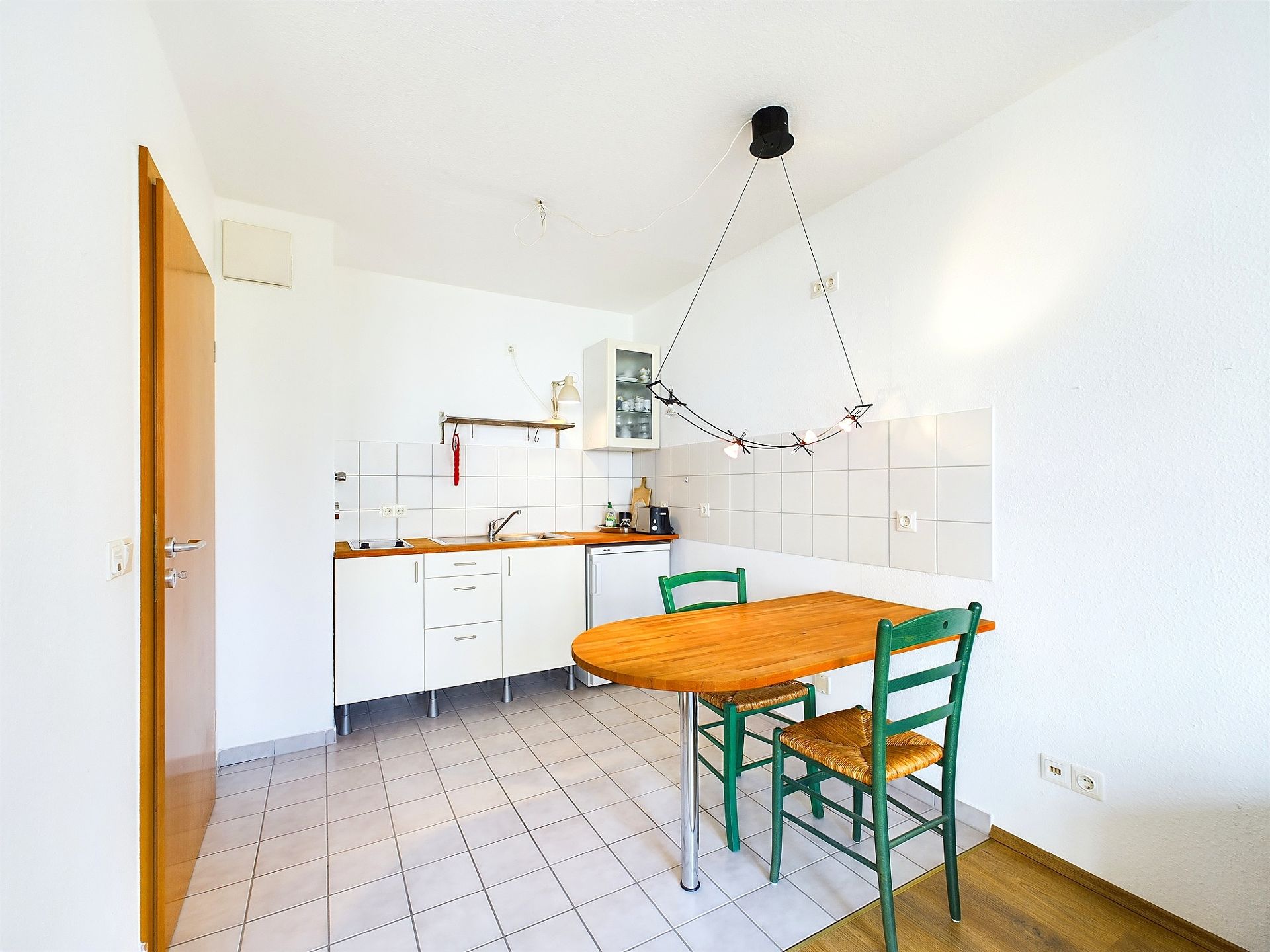 Helle Küche mit Holz-Esstisch und zwei grünen Stühlen, Wandfliesen und Hängelampe.