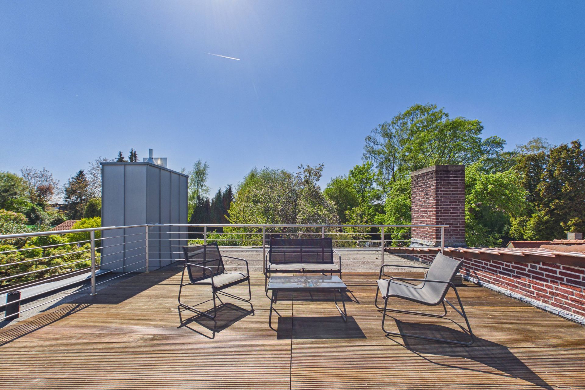 Holzterrasse mit drei Stühlen und einem Tisch, umgeben von Bäumen und blauer Himmel.