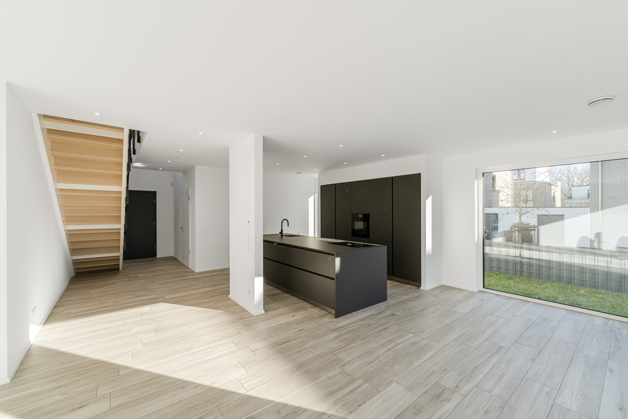 Helles, modernes Wohnzimmer mit Küche, Holztreppe und großem Fenster zum Garten.