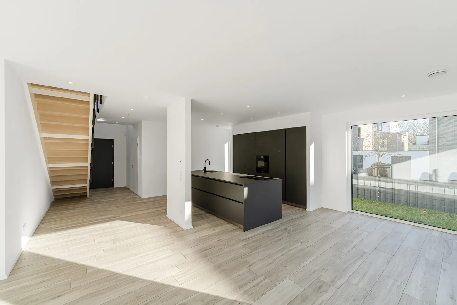 Helles, modernes Wohnzimmer mit Küche, Holztreppe und großem Fenster zum Garten.