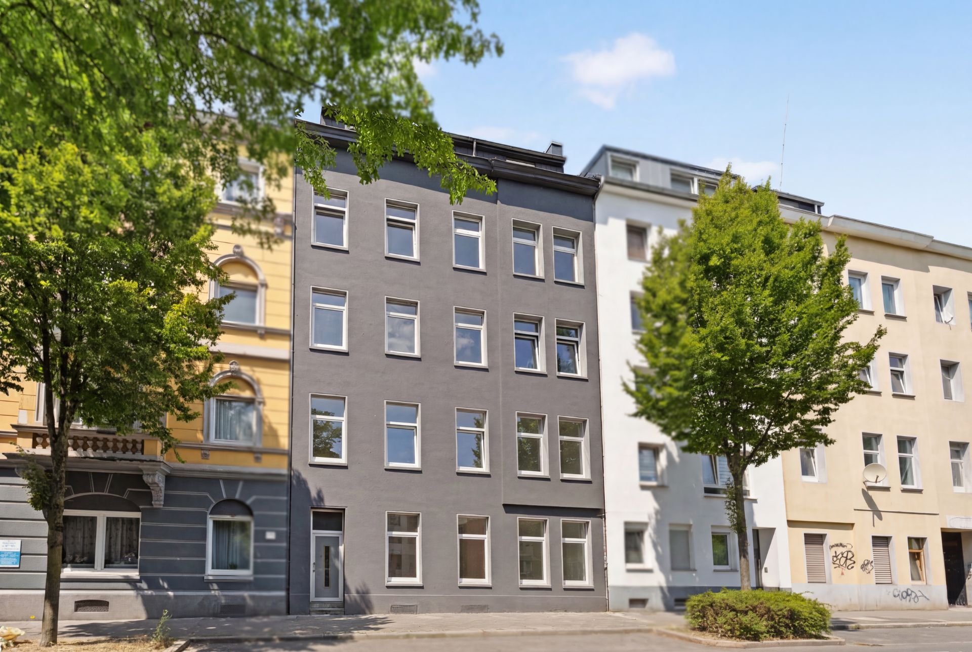 Mehrstöckiges Wohnhaus mit grauer Fassade und vielen Fenstern, Bäume stehen davor.