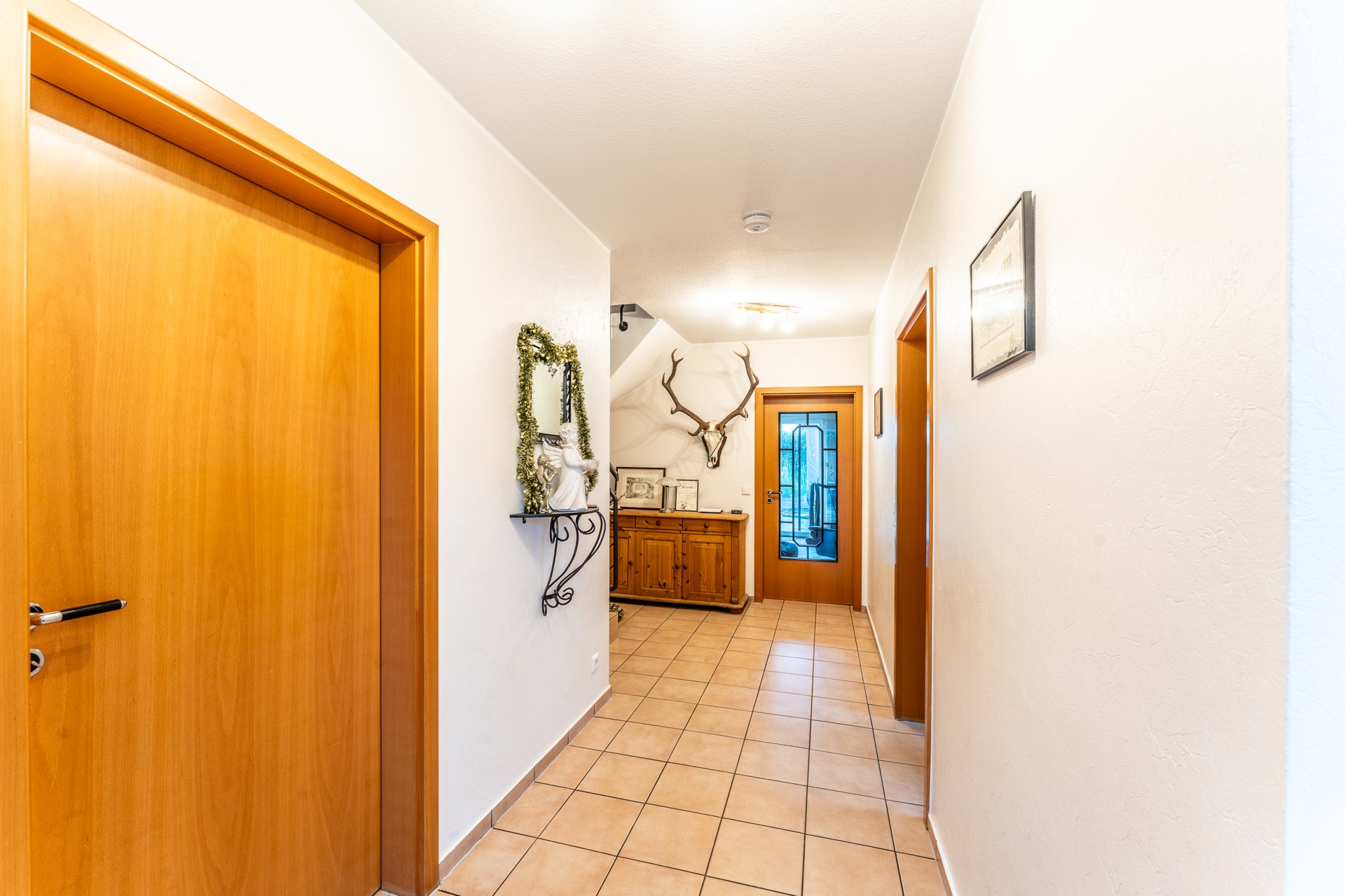 Heller Flur mit Fliesenboden, Holztüren, Sideboard, Spiegel und Hirschgeweih an der Wand.