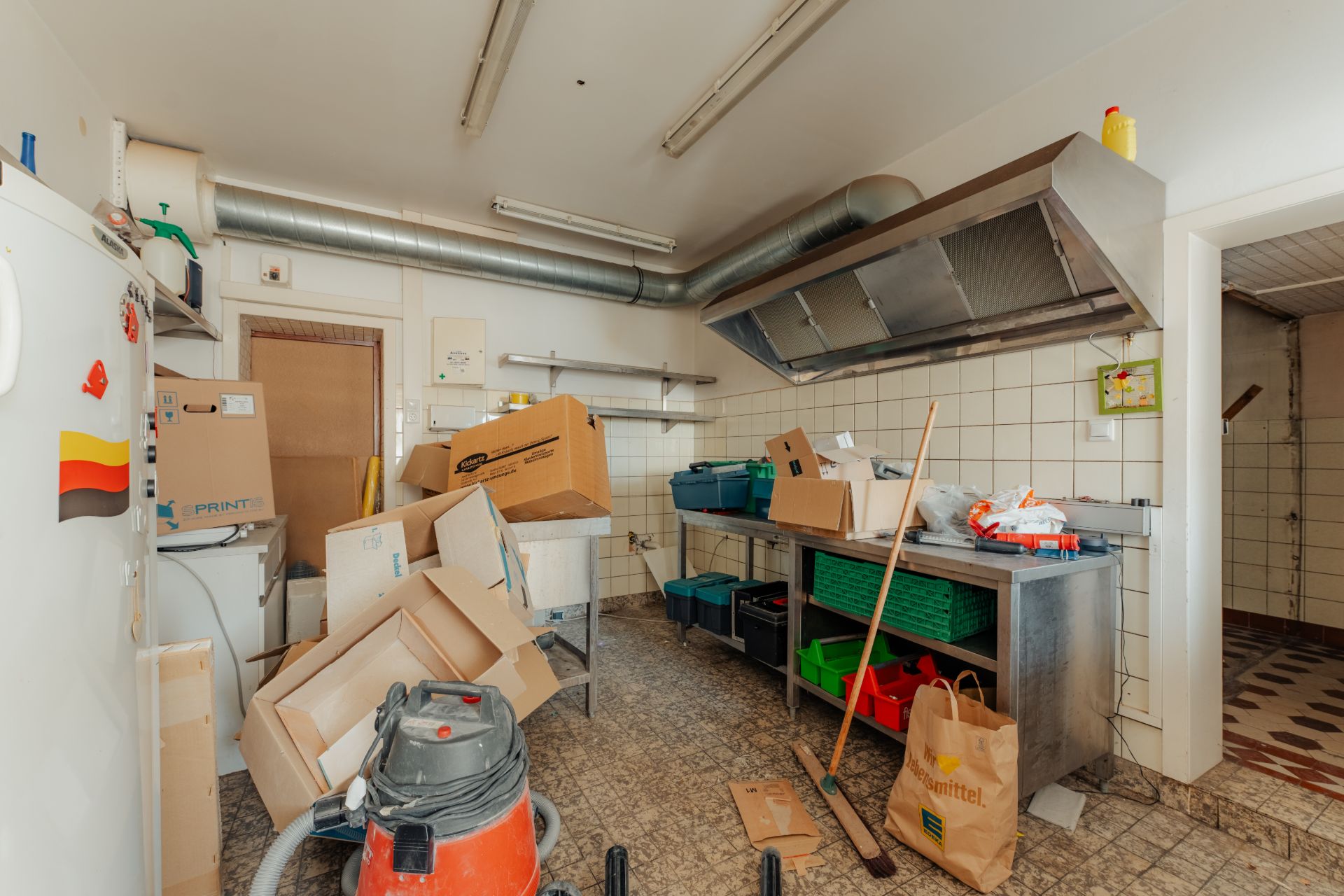 Unordentliche Küche mit Kartons, Staubsauger und Reinigungsutensilien auf Fliesenboden.