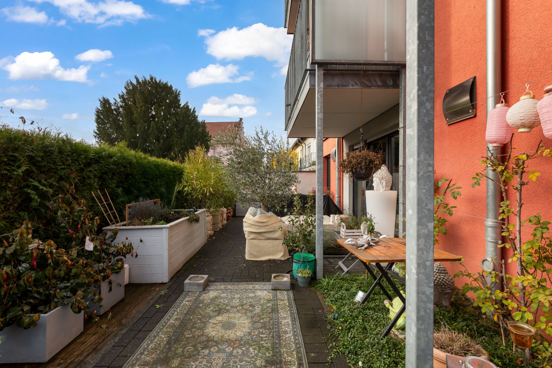 Terrasse mit Teppich, Sitzsack, Tisch und Pflanzen vor orangener Hauswand.