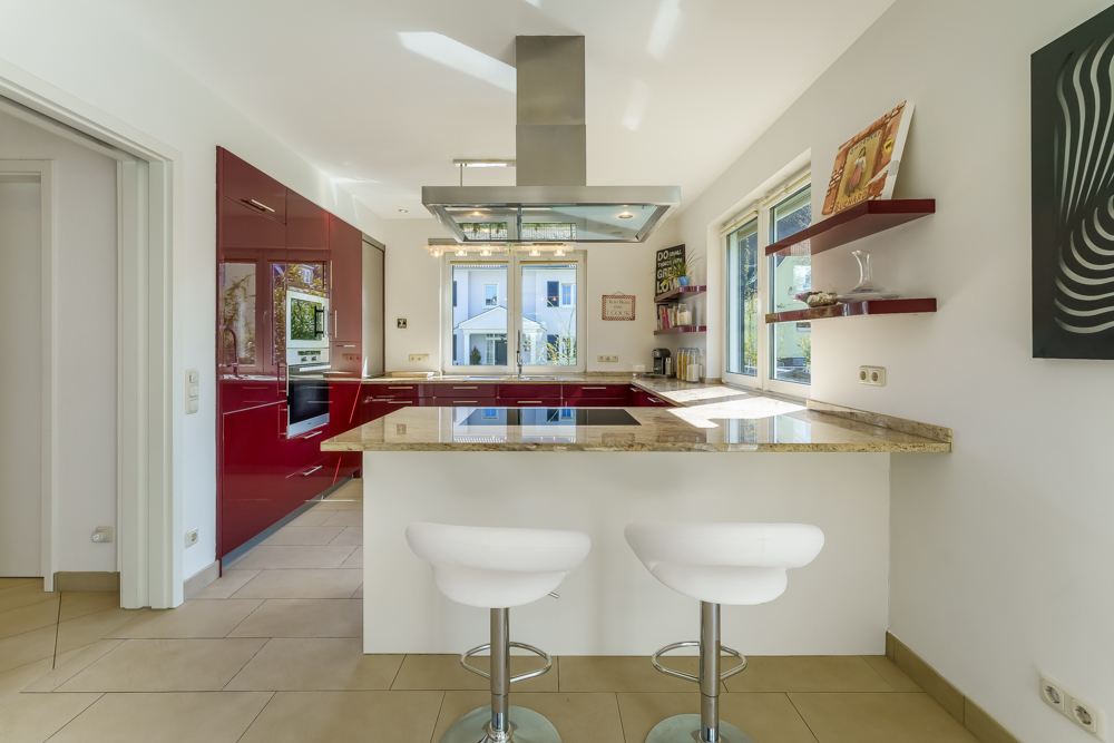 Moderne Küche mit roten Schränken, großem Fenster, Frühstückstheke und zwei weißen Barhockern.