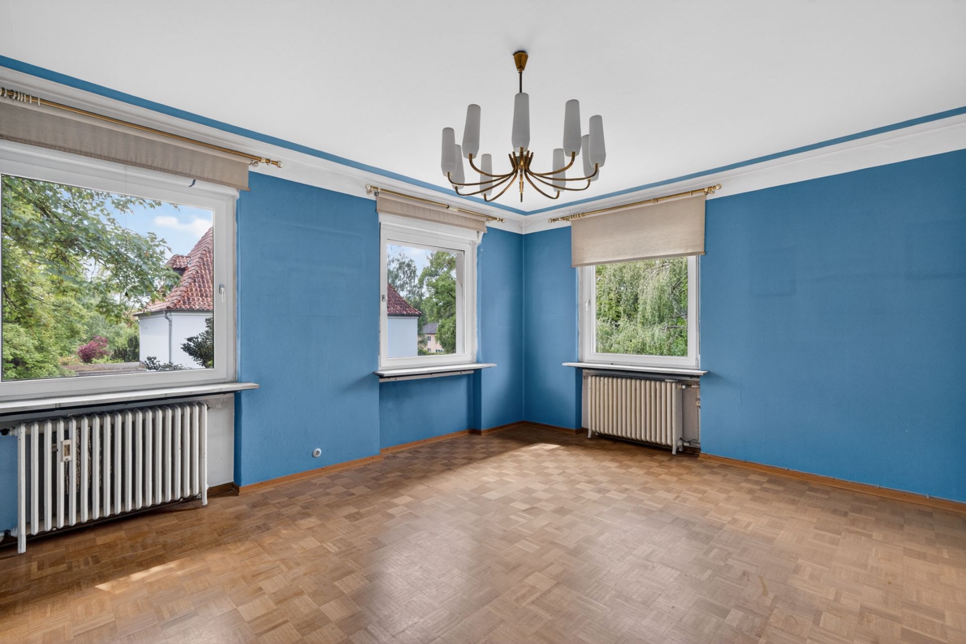 Leerer Raum mit blauen Wänden, Parkettboden, zwei Fenstern und einem Kronleuchter.