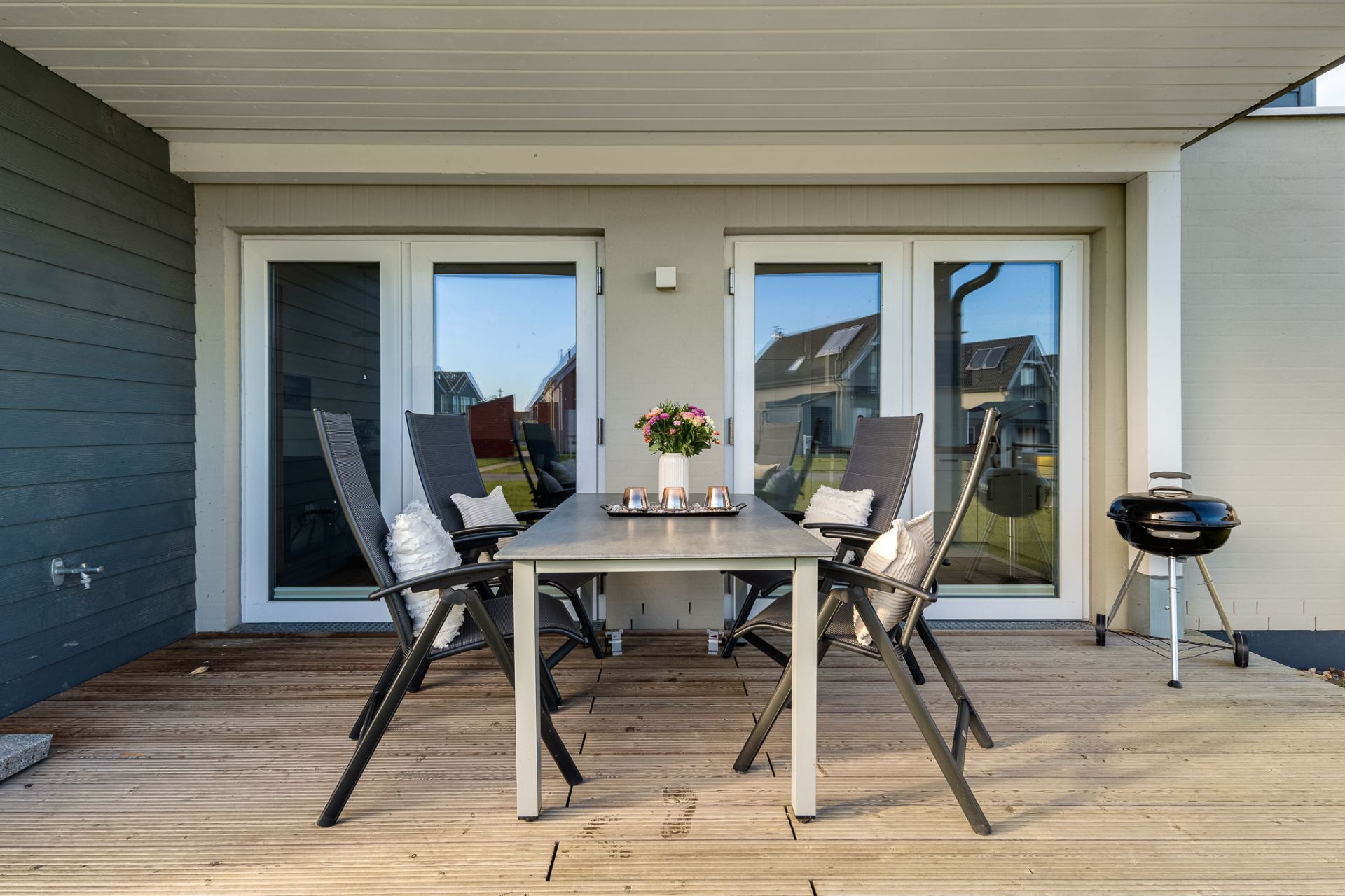 Terrasse mit Esstisch, vier Stühlen, Blumenvase und Grill vor zwei Glastüren.