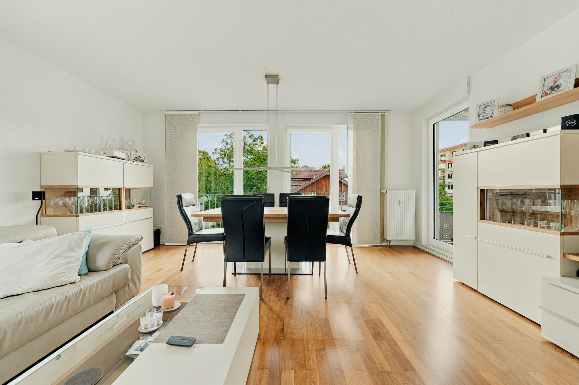 Helles Wohnzimmer mit Essbereich, Holzboden, beigen Möbeln und großen Fenstern.