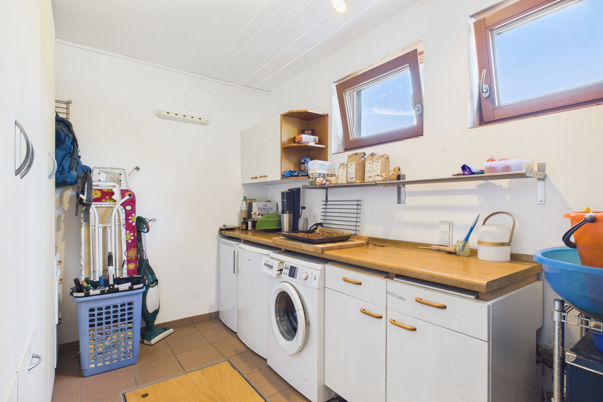 Hauswirtschaftsraum mit Waschmaschine, Arbeitsplatte, Regalen und zwei Fenstern.