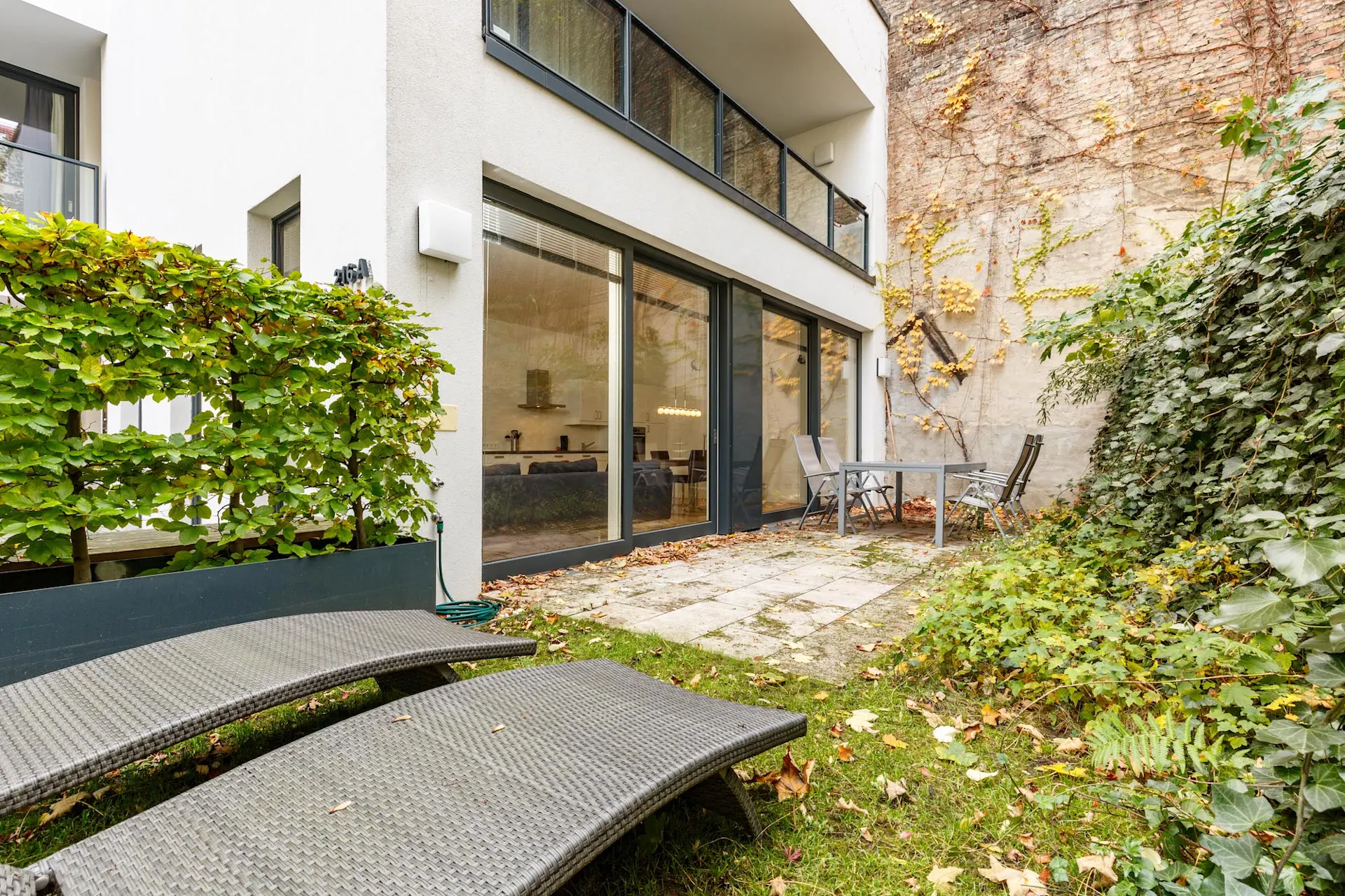 Kleine Terrasse mit Tisch, vier Stühlen und zwei Liegen neben einer modernen Hausfassade.