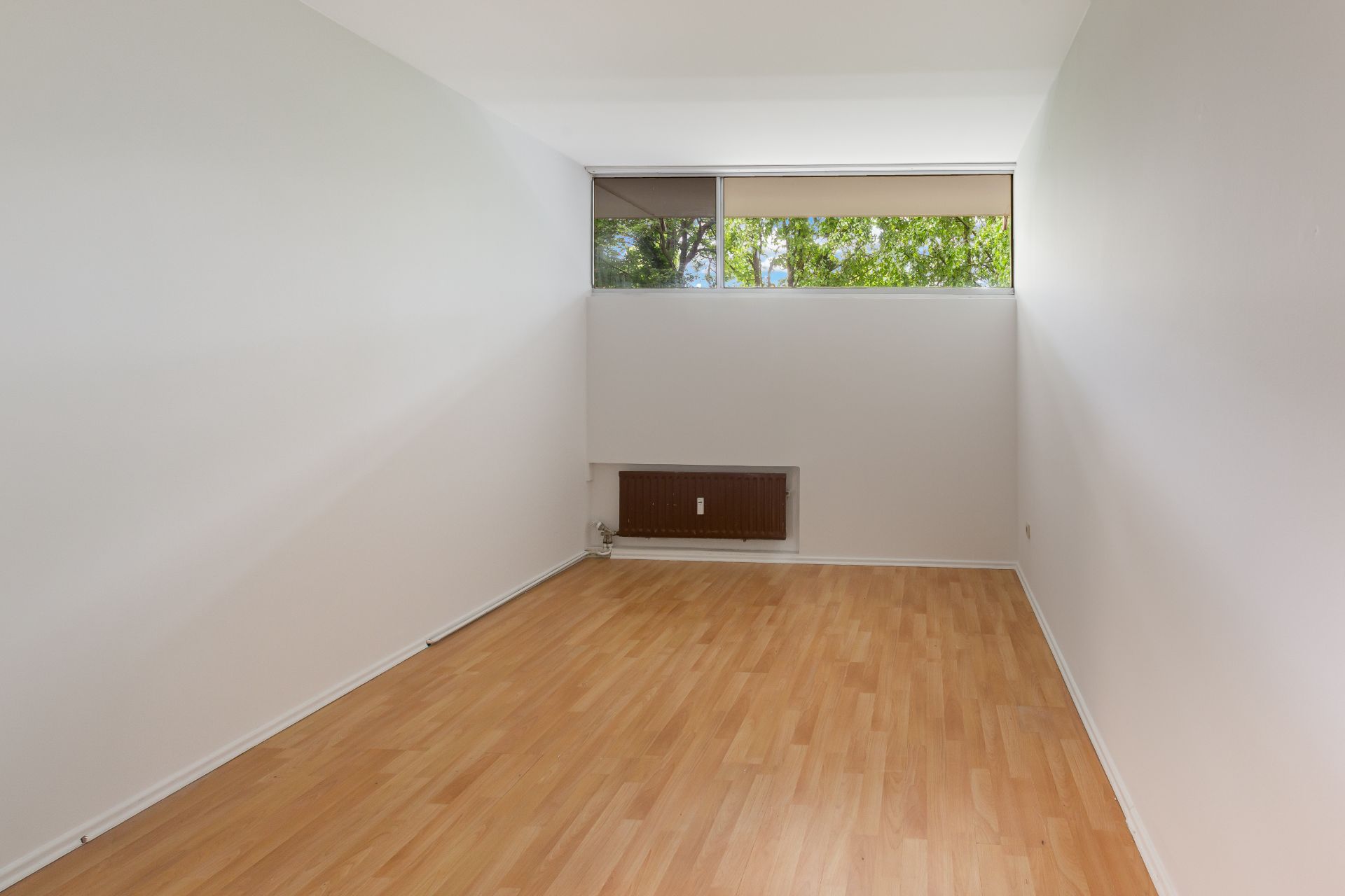 Leerer Raum mit hellem Holzfußboden, weißen Wänden und einem Fenster mit Blick auf Bäume.