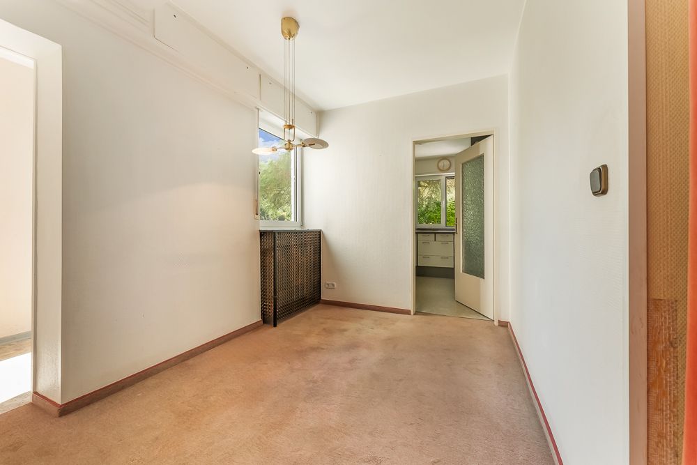 Leerer, heller Raum mit Fenster, Deckenlampe und offenem Durchgang zur Küche.