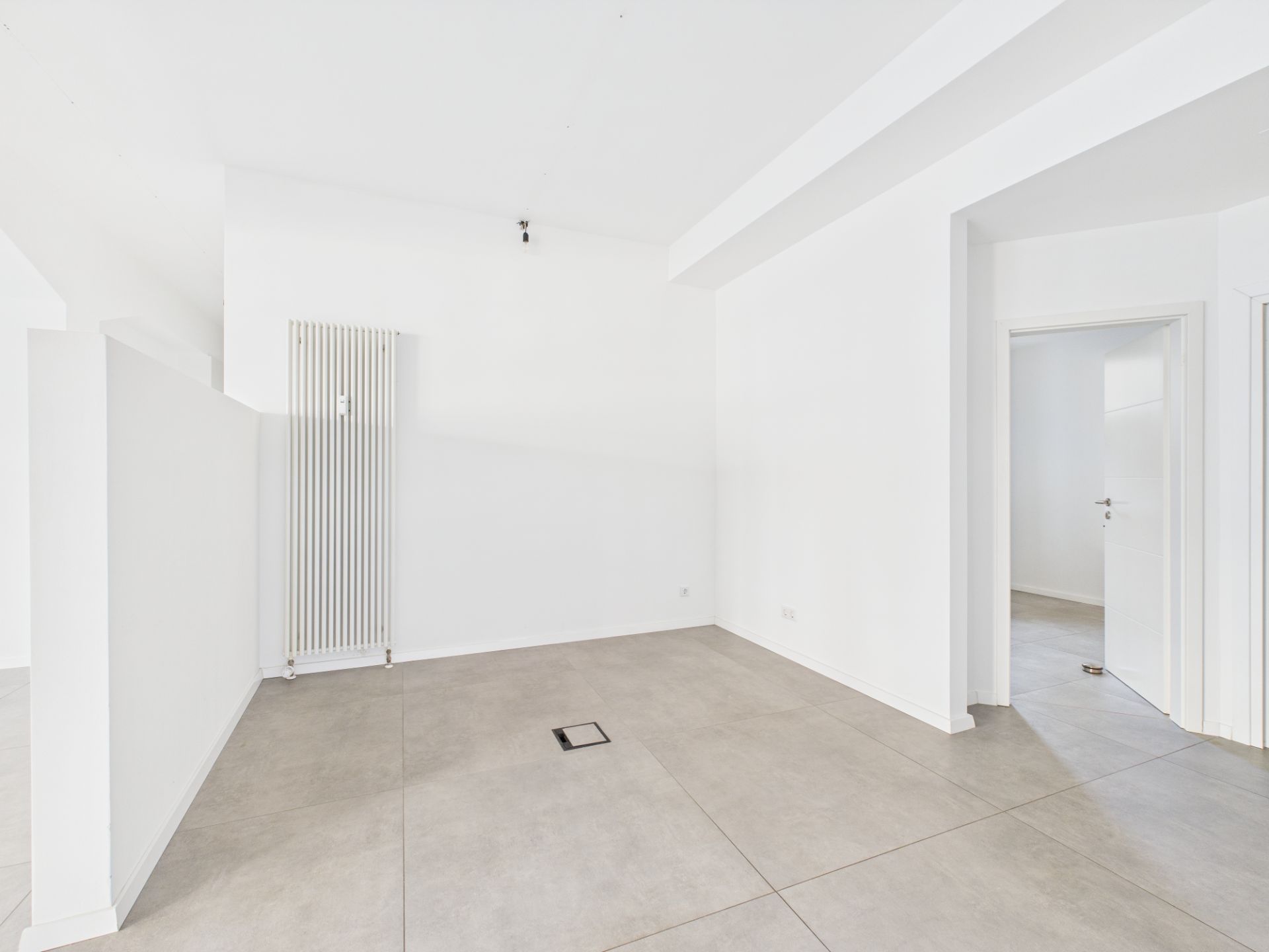 Heller, leerer Raum mit großen, grauen Fliesen und einem weißen Heizkörper an der Wand.