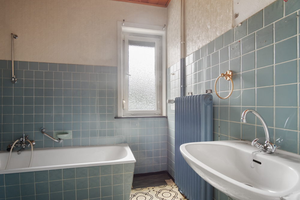 Badezimmer mit Badewanne, Waschbecken, blauem Heizkörper und Fenster, türkisfarbene Wandfliesen.