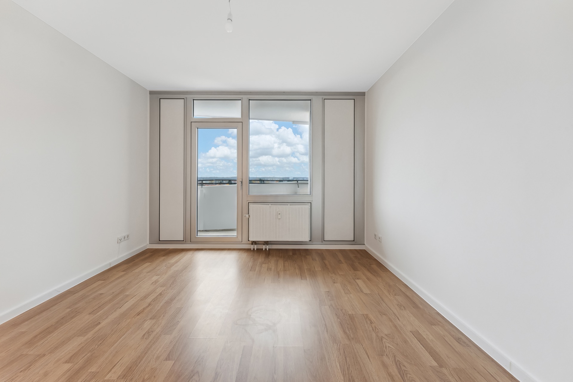 Helles, leeres Zimmer mit Holzboden, großen Fenstern und Zugang zu einem Balkon.