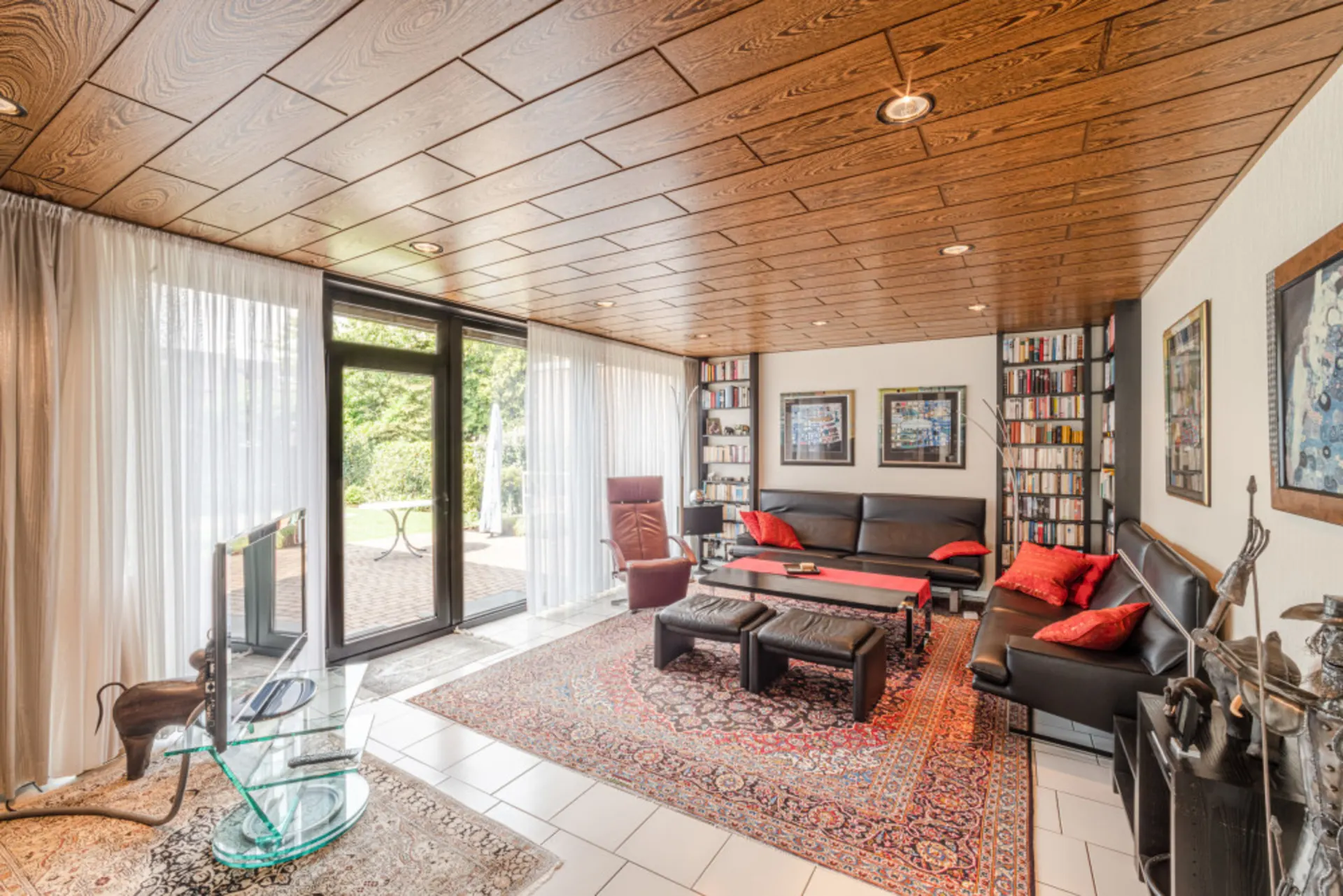 Wohnzimmer mit Ledersofas, Teppich, Bücherregalen, Glastür zum Garten und Holzdecke.