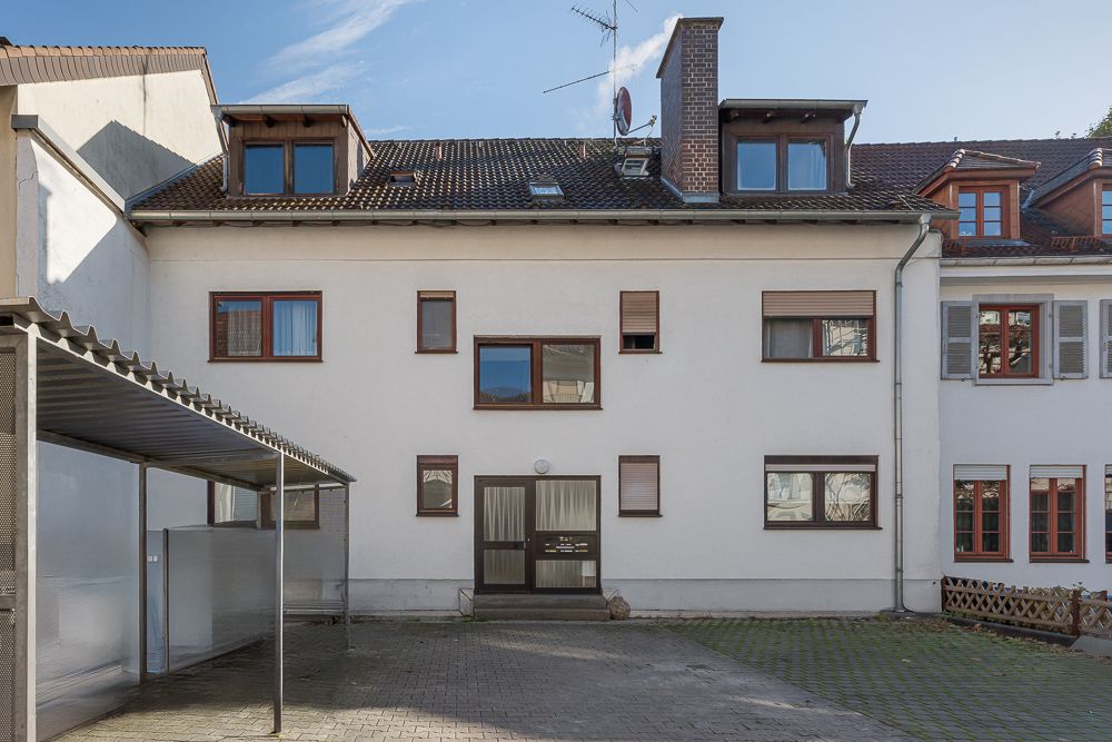 Dreistöckiges Wohnhaus mit mehreren Fenstern und einem Eingangsbereich, davor gepflasterter Hof.