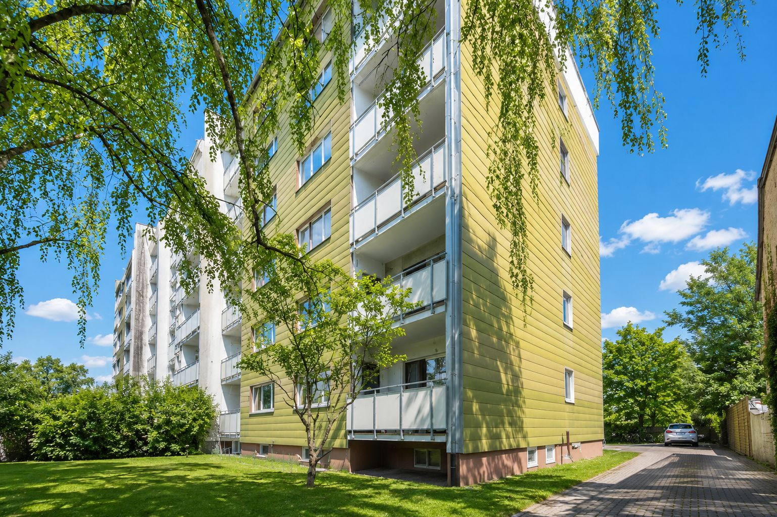 Mehrstöckiges Wohnhaus mit gelber Fassade, Balkonen und umgeben von Bäumen und grüner Wiese.