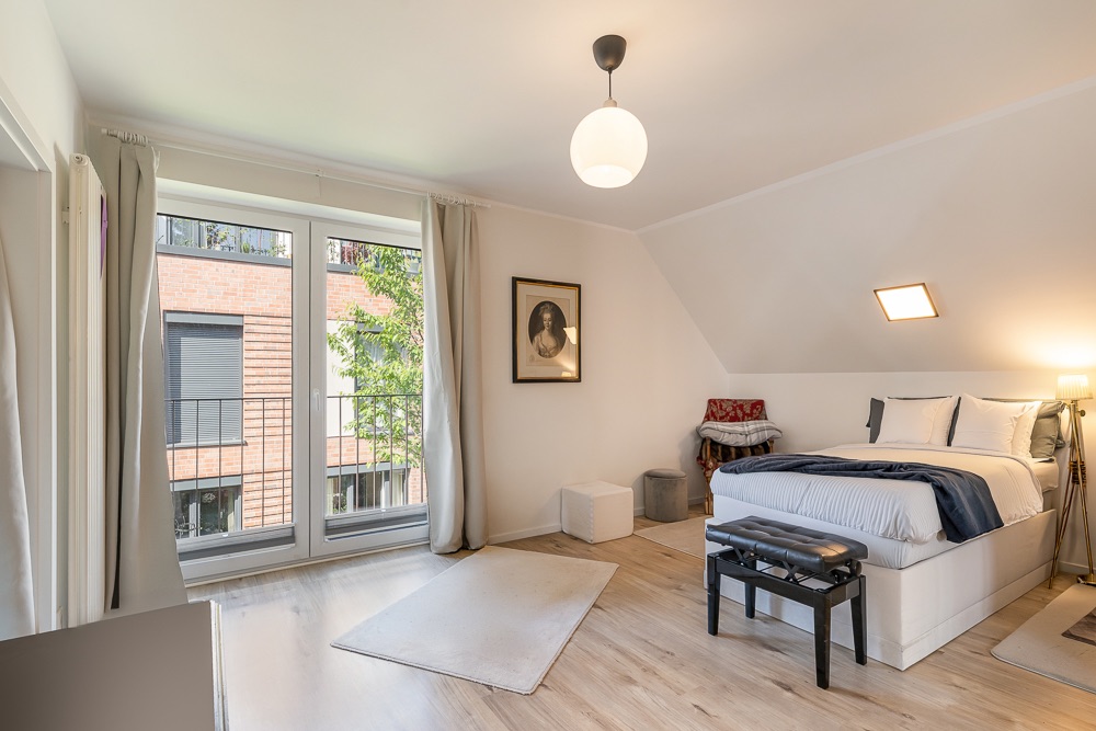 Helles Schlafzimmer mit großem Fenster, Doppelbett, hellen Vorhängen und Holzfußboden.