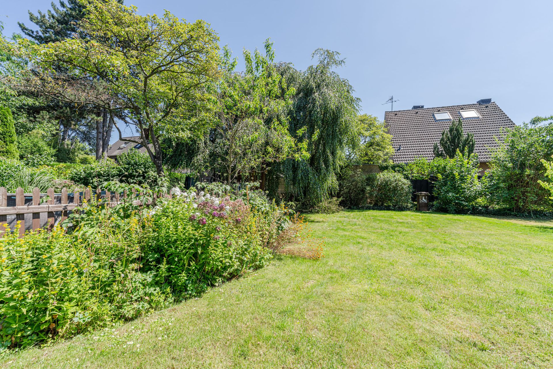 Grüner Garten mit Rasenfläche, Blumenbeeten, Bäumen und Holzzaun, Haus im Hintergrund.