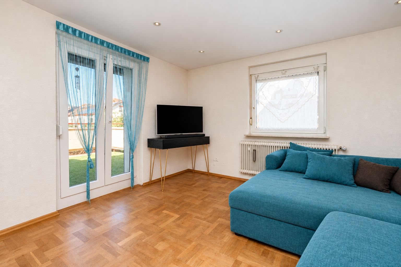 Wohnzimmer mit Parkettboden, blauer Eckcouch, Fernseher und großen Fenstern mit Vorhängen.
