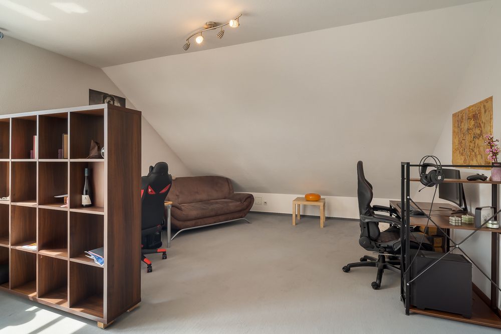 Helles Zimmer mit Regal, Schreibtisch, Bürostuhl und braunem Sofa unter einer Dachschräge.