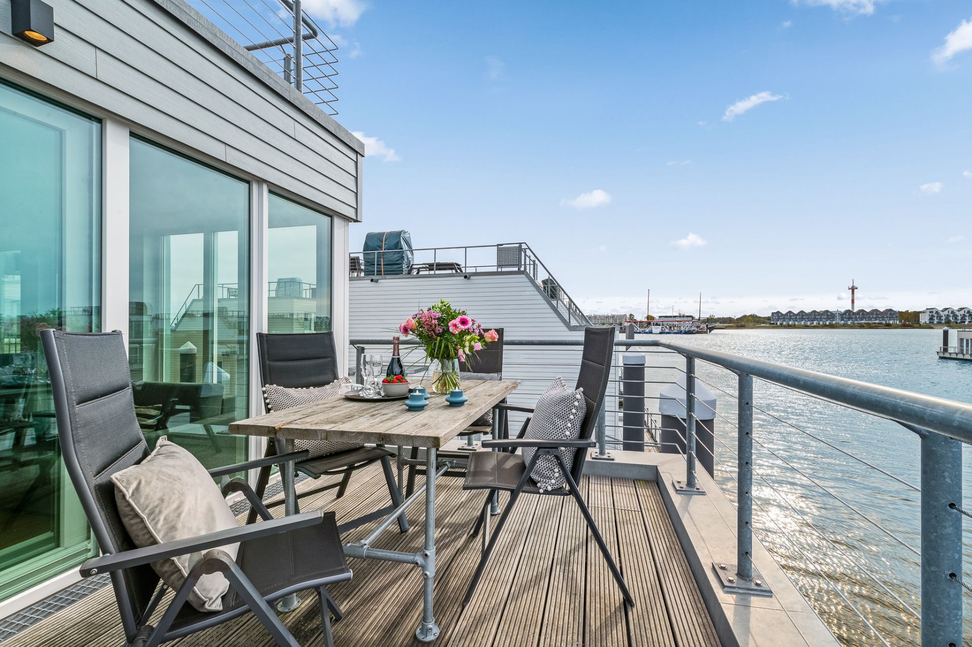 Sonnige Terrasse mit Holzboden, Tisch, vier Stühlen und Blick auf das Wasser.
