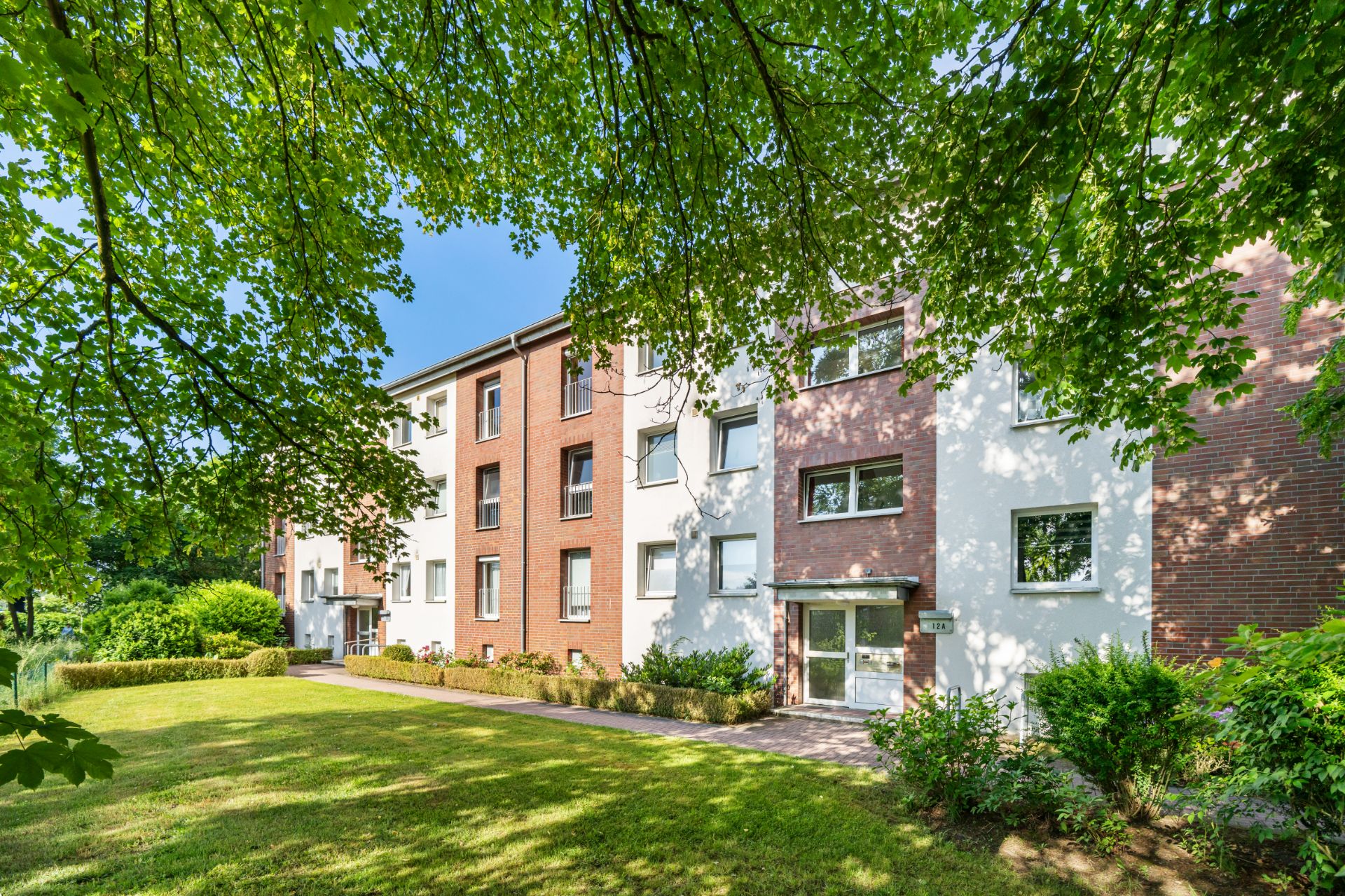 Mehrfamilienhaus mit drei Stockwerken und Vorgarten, umgeben von Bäumen.