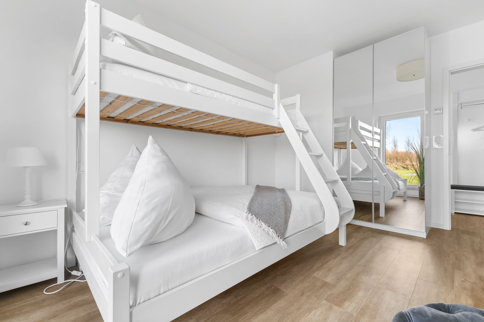 Helles Schlafzimmer mit weißem Etagenbett, Nachttisch und Spiegelschrank auf Holzfußboden.