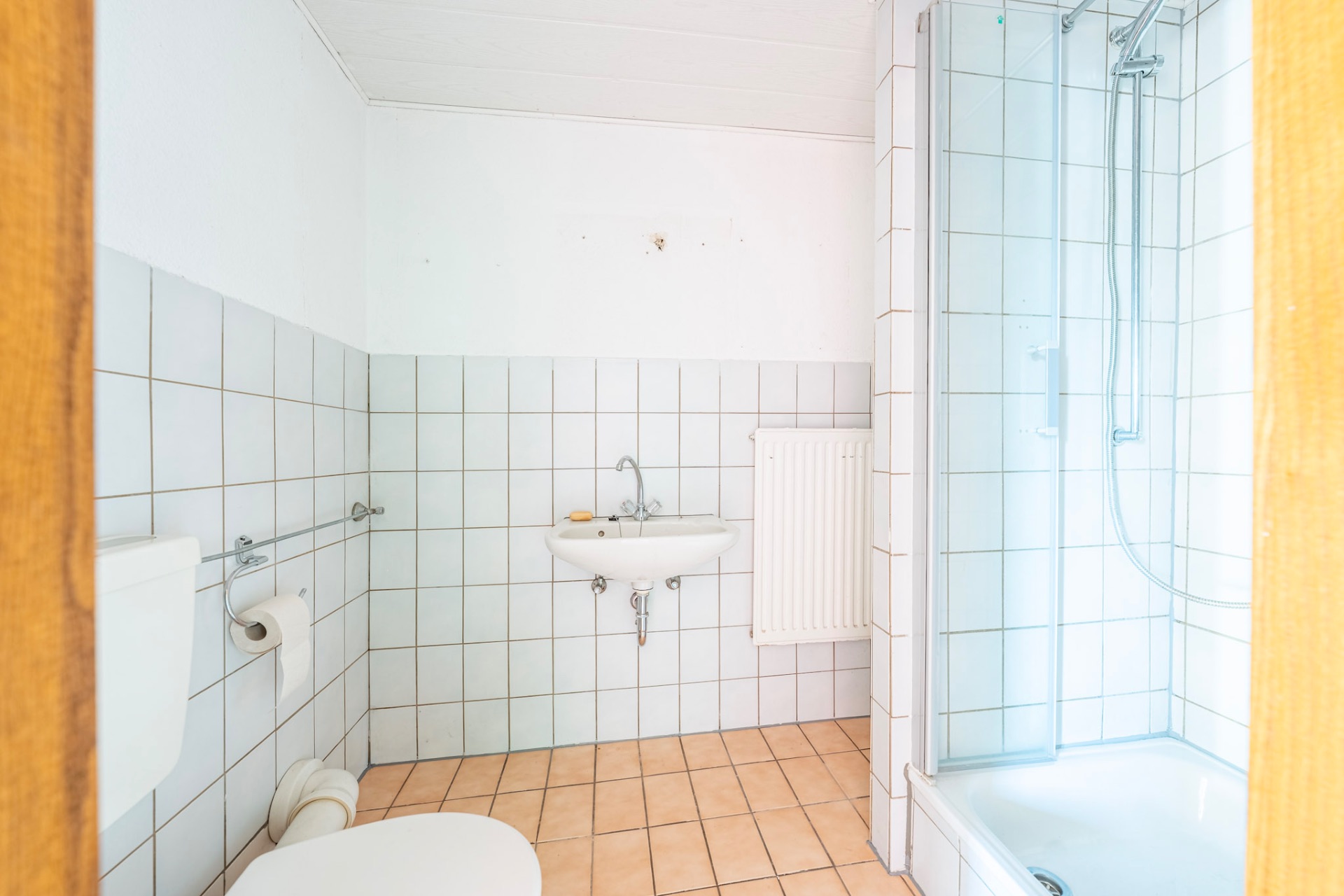 Helles Badezimmer mit Toilette, Waschbecken, Dusche und Heizkörper, gefliest an Wänden und Boden.