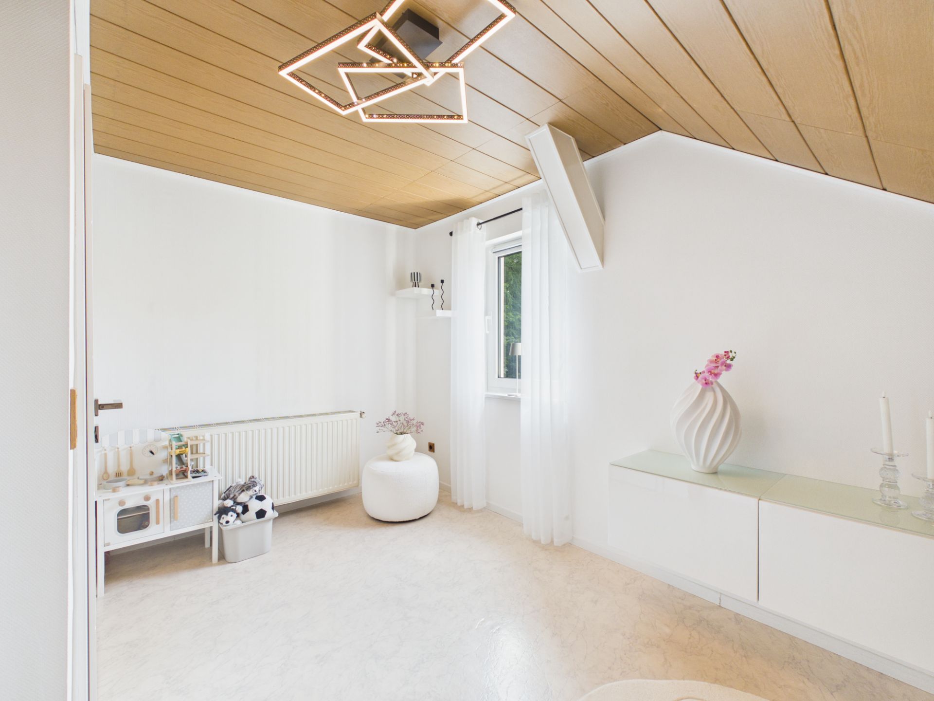 Helles Zimmer mit Holzdecke, modernem Deckenlicht, Fenster, weißen Möbeln und Deko.