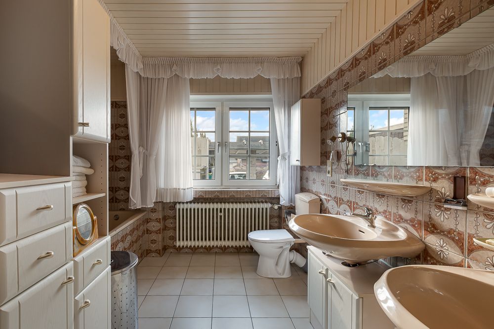 Helles Badezimmer mit zwei Waschbecken, Toilette, Spiegelschrank und großem Fenster mit weißen Vorhängen.