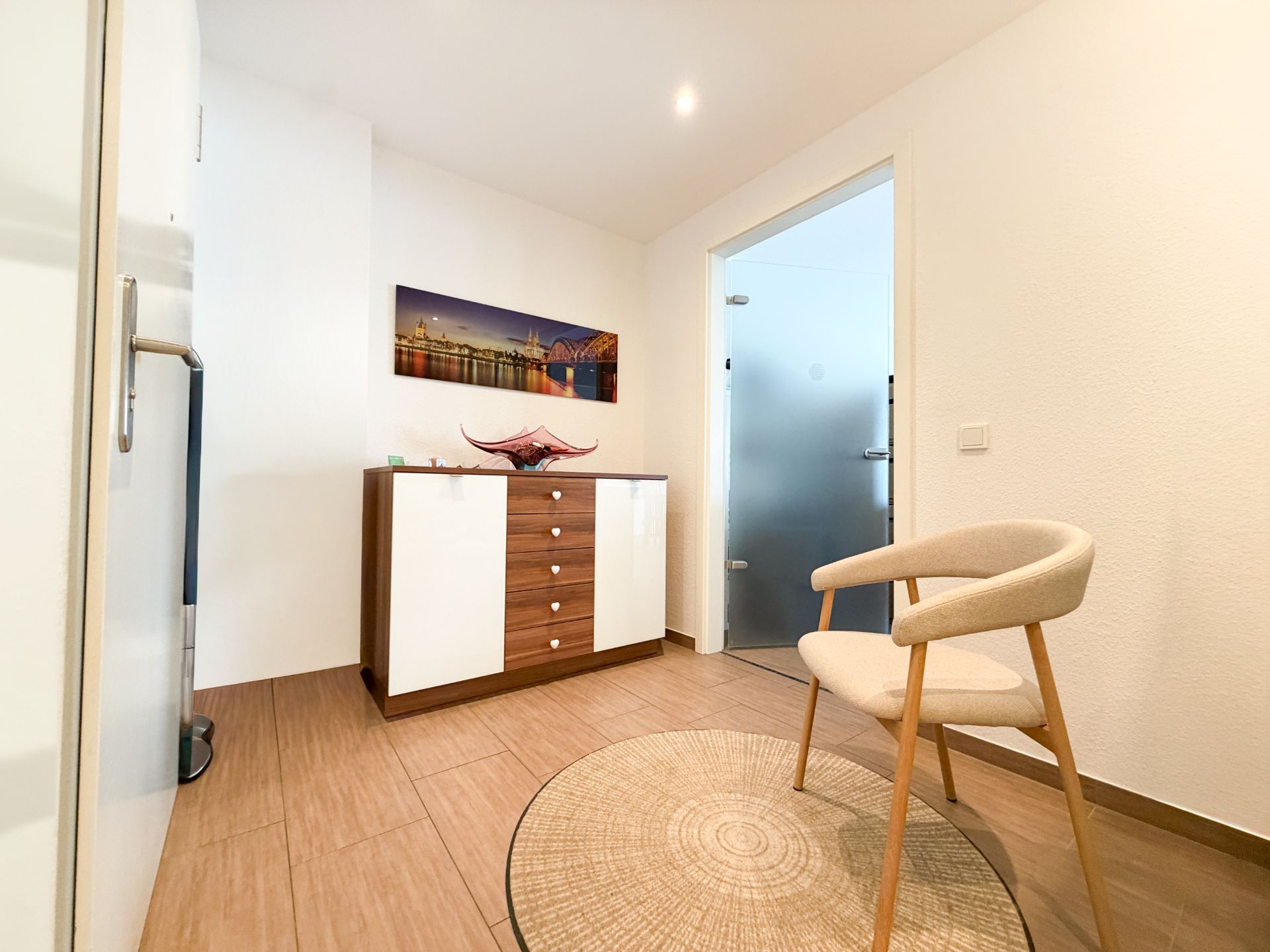 Heller Raum mit Stuhl, Teppich, Kommode und Wandbild. Tür mit Milchglas im Hintergrund.